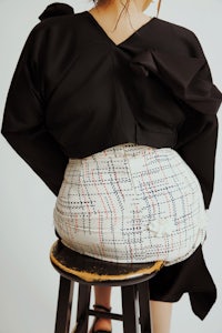 a woman sitting on a stool wearing a black top and a plaid skirt